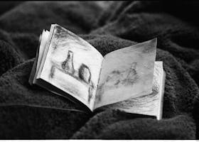 A black and white photo of an open sketchbook with abstract drawings, resting on a textured blanket.