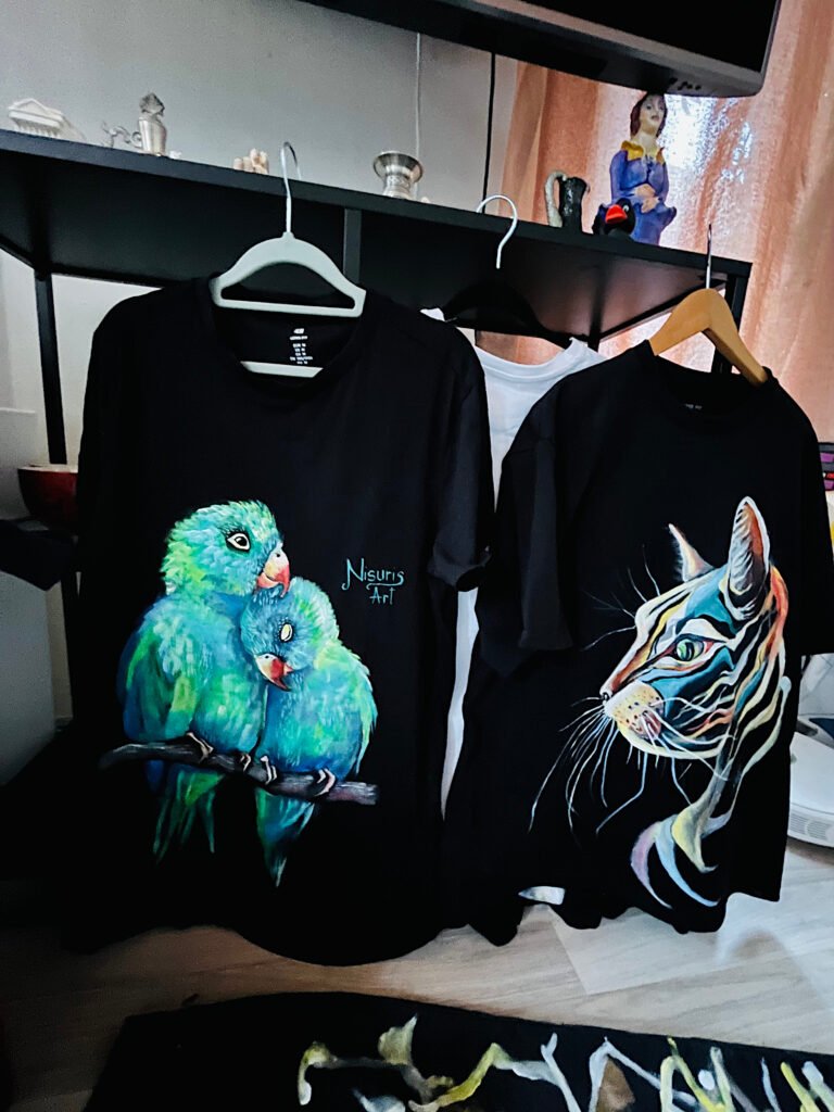 Two finished hand-painted black T-shirts with colorful parrot and cat designs